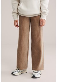 Persona che indossa pantaloni beige a coste a gamba larga, felpa bianca e sneakers argento-bianco in piedi su uno sfondo neutro.