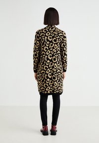 Lång svart cardigan med leopardmönster i nyanser av beige, med långa ärmar och ribbat avslut. Bärs över svarta byxor och skor.