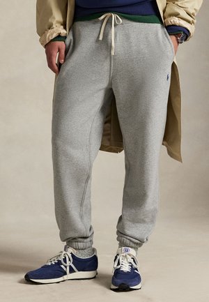 Homme portant un pantalon de jogging gris à cordon, des baskets bleues, des chaussettes grises, un trench-coat beige et un haut bleu, debout les mains dans les poches.