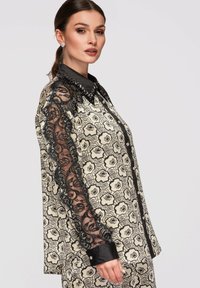 Femme portant une chemise à manches longues avec boutons, manches en dentelle noire et tissu crème orné de motifs floraux noirs, posant de profil.