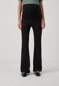 Pantalons de maternité noirs évasés avec une taille élastique, texture détaillée et une finition lisse. Le modèle porte des chaussures noires.