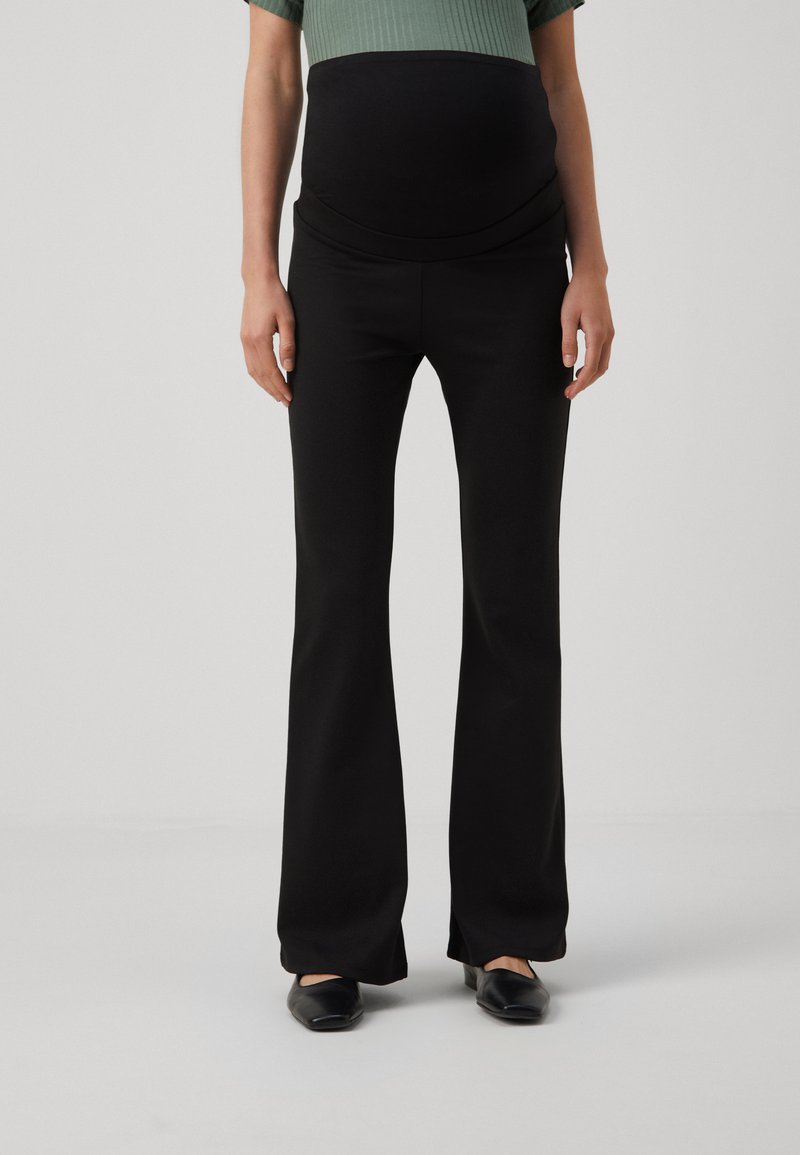 Pantalons de maternité noirs évasés avec une taille élastique, texture détaillée et une finition lisse. Le modèle porte des chaussures noires.