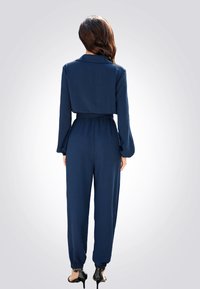 Combinaison bleu marine avec un col, des manches longues, une taille élastique et des chevilles fuselées. Texture lisse et silhouette ajustée.
