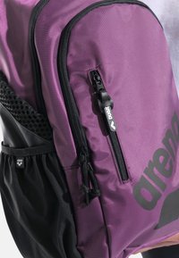 Zaino in tessuto viola con dettagli neri, dotato di una cerniera ampia, tasca laterale in rete e un grande logo sul davanti. Design compatto.