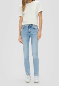 Femme portant une chemise blanche à manches courtes, un jean slim bleu clair et des baskets blanches, posant devant un fond clair uni.