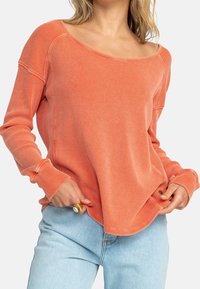 Haut à manches longues et épaules dénudées de couleur corail texturée, avec tissu côtelé et détails cousus. Associé à un jean délavé clair.