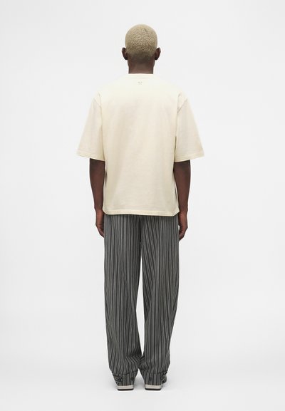 Άτομο με κοντά ξανθά μαλλιά που φοράει μία φαρδιά μπεζ μπλούζα και χαλαρό γκρι παντελόνι με ρίγες, στέκεται με το πρόσωπο προς ένα απλό, ανοιχτό φόντο.