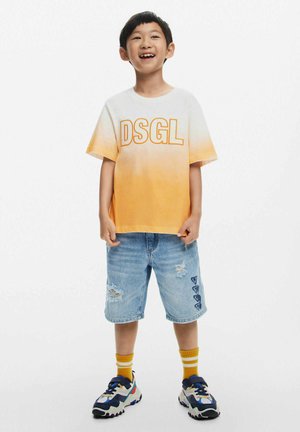 Camiseta degradada en naranja con el texto "DSGL", combinada con pantalones cortos de mezclilla azul desgastados y zapatillas coloridas. Fondo blanco.