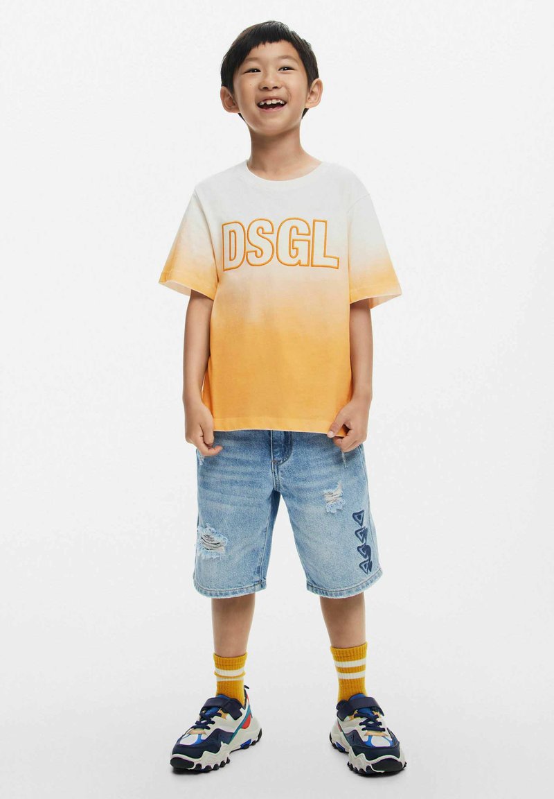 T-shirt con gradiente arancione e testo "DSGL", abbinato a pantaloni shorts in denim blu sfilacciati e sneakers colorate. Sfondo bianco.