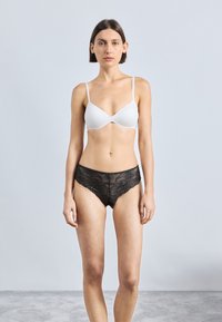 Woman standing facing forward wearing a white bra and black lace underwear against a plain light grey background.