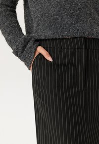 Pull en tricot gris foncé à manches longues associé à un pantalon noir à rayures verticales avec un devant plat et une coupe élégante, ajustée.