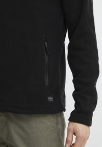 Sort fleece-pullover med en sidelomme med lynlås, tekstureret overflade og et lille logo mærke i kanten. Har lange ærmer og lige pasform.