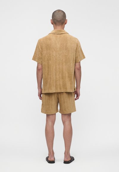 Homme vu de dos portant une chemise beige à manches courtes et un short assorti en tissu texturé avec des sandales noires.