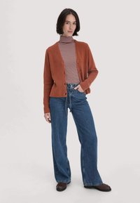 Vrouw die staat en een bruine gebuttonde cardigan, gestreepte coltrui, wijdeloze blauwe jeans en donkerbruine schoenen draagt tegen een effen achtergrond.