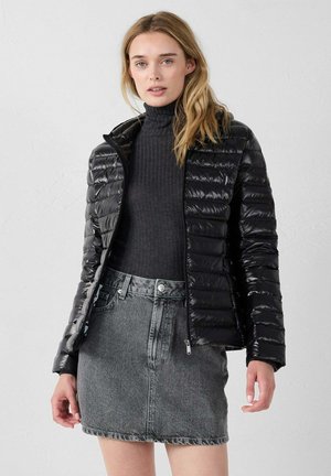 Mujer con una chaqueta acolchada negra sobre un suéter de cuello alto negro y una falda de mezclilla gris, de pie frente a una pared blanca lisa.