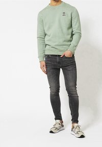Felpa verde menta con polsini e orlo a costine, abbinata a jeans slim-fit grigio scuro e sneakers beige con motivo camouflage.