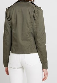 Olivově zelená zkrácená pracovní bunda s ramenními epauletami, texturovaná látka a rovný střih. Nosená přes bílé kalhoty.