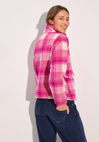 Veste en polaire rose et blanche à carreaux avec un col, deux poches et une coupe décontractée. Portée avec un pantalon bleu foncé, vue de dos.
