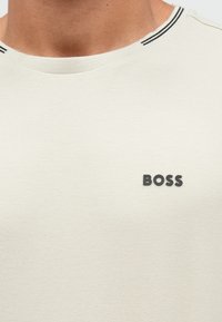 Ανοιχτό μπεζ πόλο με υφή, με σκούρο γκρι λογότυπο "BOSS" και γιακά με ασπρόμαυρες ρίγες.