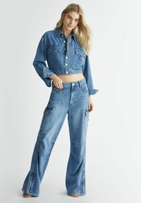 Giacca di jeans tagliata con due tasche frontali, colore azzurro chiaro, chiusura con bottoni, abbinata a jeans cargo a vestibilità ampia, con tasche laterali.