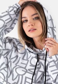 Biała bluza z kapturem z czarnym geometrycznym wzorem, wyposażona w sznurki i luźny krój. Miękka tkanina o gładkiej fakturze.