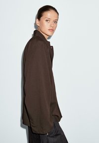 Bruine lange mouwen shirt met hoge kraag, losse pasvorm en textuurstof. Heeft een enkele knoopdetail bij de hals.