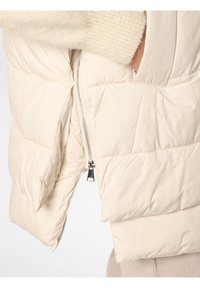 Krémová péřová vesta s prošívaným designem, která má boční zip a hladkou strukturu. Vesta má zakulacený střih na spodním okraji.