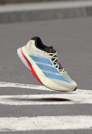 Chaussure de course légère blanche et bleue avec un détail de semelle rouge flottant au-dessus d'une surface pavée avec des lignes blanches peintes.