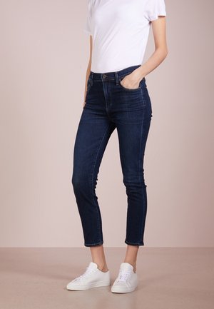 Vaqueros skinny de mezclilla azul oscuro con cintura alta, que presentan bolsillos frontales y una longitud recortada, combinados con zapatillas blancas y una camiseta blanca básica.