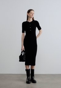 Μαύρο πλεκτό φόρεμα με κοντά μανίκια, λεπτομέρεια με κουμπιά, εφαρμοστό, μήκος μέχρι το γόνατο, συνδυασμένο με μαύρες ψηλές μπότες και μικρή τσάντα χειρός. Μινιμαλιστικός σχεδιασμός.