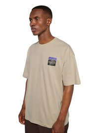 Mężczyzna z krótkimi włosami i kolczykiem, ubrany w beżowy t-shirt z niebiesko-czarnym logo „HALO” na piersi, stojący na białym tle.