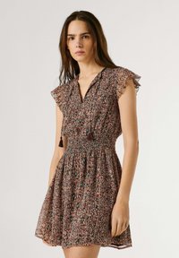 Vrouw draagt een korte, patroonjurk met wappermouwen en een geplooide taille, staand tegen een effen lichte achtergrond.