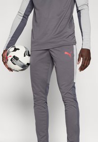 Γκρι αθλητικό παντελόνι με ανοιχτό γκρι πλαϊνές ρίγες, που φέρει κόκκινο λογότυπο Puma. Το άτομο κρατά μια ασπρόμαυρη μπάλα ποδοσφαίρου.