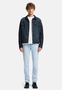 Kurtka dżinsowa w ciemnoniebieskich i jasnoniebieskich odcieniach, z zapięciem na guziki z przodu, dwoma kieszeniami na piersi, noszona na białej koszuli, w komplecie z jasnoniebieskimi dżinsami i czarnymi butami.