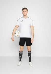 Witte sportieve shirt met korte mouwen met zwarte strepen, zwarte shorts, zwarte kniekousen met witte strepen, en witte voetbalschoenen.