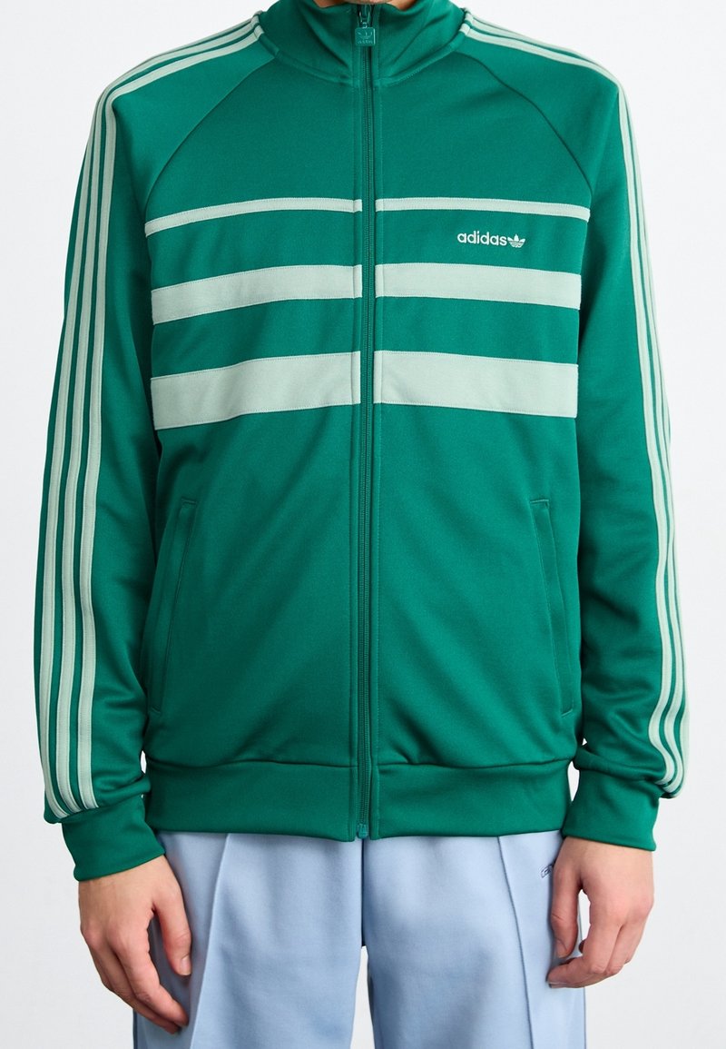 Grön Adidas-träningsjacka med tre ljusgrå horisontella ränder, frontdragkedja och ribbade ärmmuddar. Accentuerad med signaturlogotyp.