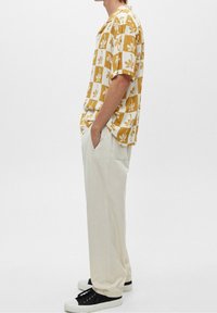 Camisa de manga corta en un patrón a cuadros amarillo y blanco con diseño de hojas, combinada con pantalones color crema y zapatillas negras.