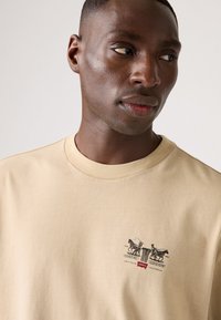 Beige t-shirt met korte mouwen, gemaakt van katoen, met een grafische print van paarden en figuren. Het ontwerp bevat tekst met de woorden "Crafted in Levi's California."