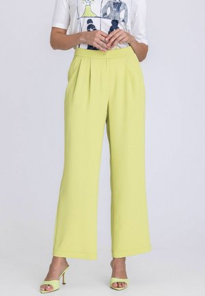 Femme portant un pantalon jaune taille haute à jambes larges, assorti à des talons ouverts jaunes et un haut blanc à manches courtes avec un motif graphique.
