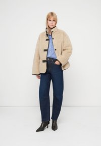 Veste polaire beige avec boutons noirs, col montant et poches latérales. Associée à une chemise rayée bleue et un jean bleu foncé. Bottines noires.
