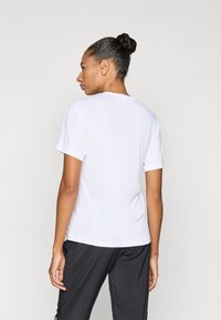 Bijela kratka majica s okruglim izrezom i opuštenim krojem, izrađena od glatkog tkanja, prikazana s leđa. Vidljive crne hlače.