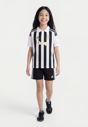 Mädchen geht vorwärts und trägt ein schwarz-weiß gestreiftes Juventus-Fußballtrikot, schwarze Adidas-Shorts und schwarze Sneaker.