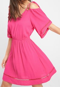 Vestido rosa con mangas cortas y hombros al aire, cintura elástica y detalles de encaje en los dobladillos de las mangas y el borde inferior. Tejido ligero.