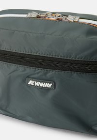 Přenosná taška K-Way v tmavě zeleném nylonu s texturovaným povrchem, která má černý zip, stříbrné kovové detaily a reliéfní logo.