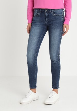 Vaqueros ajustados de mezclilla azul con un acabado desgastado, cinco bolsillos y herrajes decorativos. Combinados con una blusa rosa y zapatillas blancas.