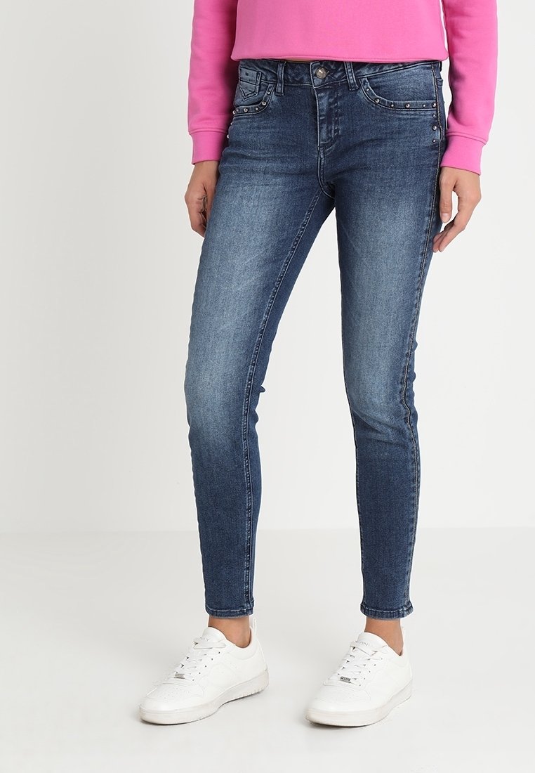 Jean denim bleu skinny avec une finition délavée, cinq poches et des accessoires décoratifs. Porté avec un haut rose et des baskets blanches.
