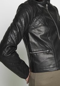 Giubbotto in pelle nera con spalle trapuntate, tasca laterale con cerniera e texture liscia, con un design attillato e colletto alto.
