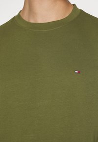 Maglietta verde oliva con scollo tondo, realizzata in tessuto morbido, con un piccolo logo ricamato in rosso, bianco e blu sul petto.