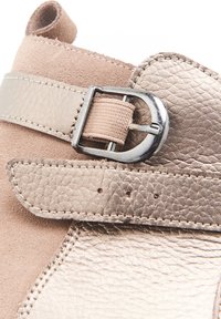 Bottine beige avec des matériaux en daim et cuir texturé. Elle présente une boucle et une sangle métalliques avec des coutures décoratives et des perforations.