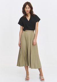 Czarna bluzka z dekoltem w serek i krótkimi rękawami, połączona z lekko opadającą, plisowaną spódnicą midi w kolorze khaki. Modelka nosi białe sandały na ramiączkach.